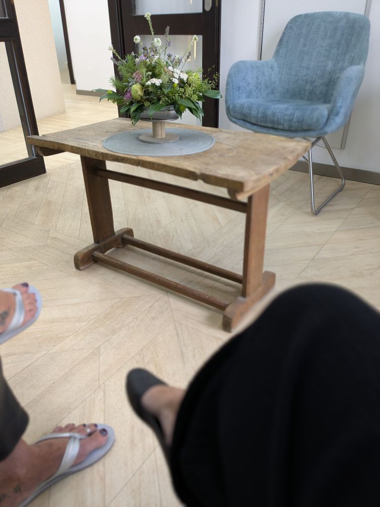 Ein Wartezimmerfußboden mit zwei Fußpaaren. In der Mitte ist auf einem alten Holzstisch ein Blumenbouquet zu sehen