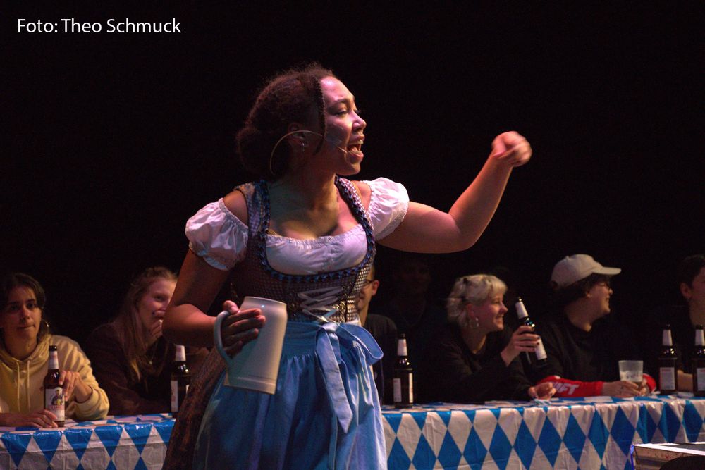 Eine Frau im Dirndl hat einen Bierkrug in der Hand und gestikuliert. Im Hintergrund stehen Biertische, an denen Bier trinkende Personen sitzen.
