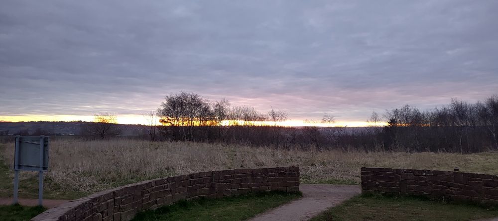 Early morning cloudy view from on top a grassy hill with a stone circle at the peak, looking out towards the sunrise on the horizon