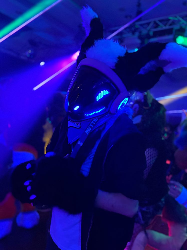 A protogen fursuiter dancing with different coloured lights around them