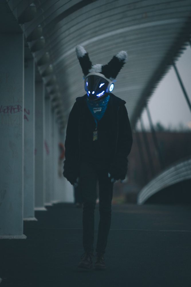 A protogen fursuiter walking along a bridge towards the camera.
The picture has muted and darker colours.
The protogen's blue face lights stand out from the darker background .