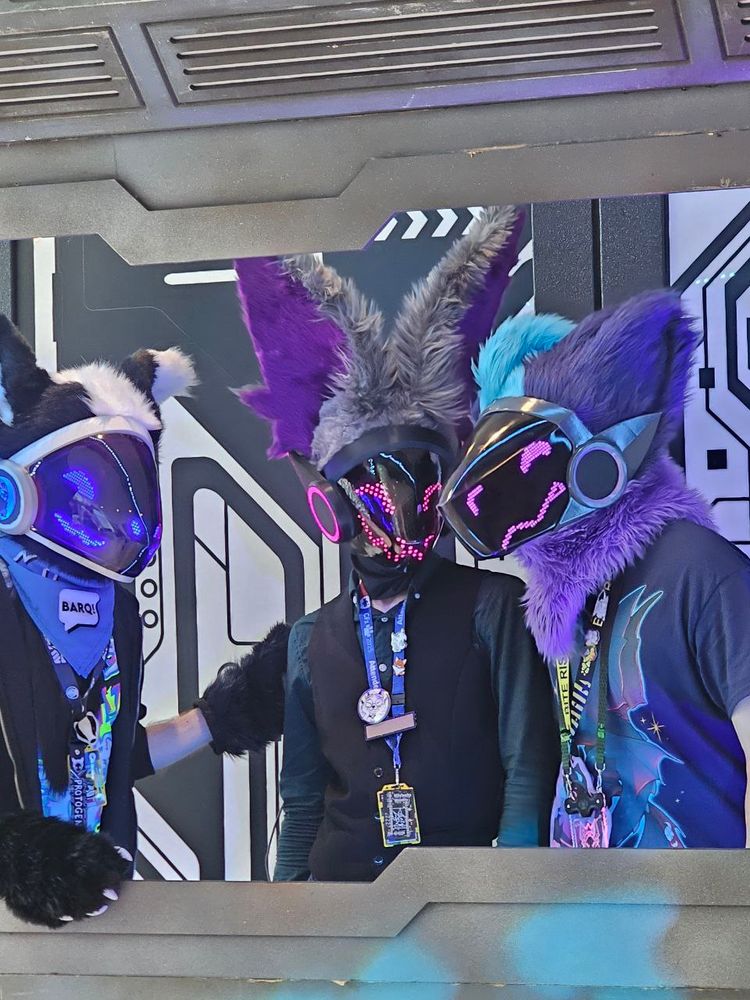 Three protogen fursuiters posing for a picture.
One with blue lights, with black and white fur.
One with pink/purple lights, with grey and purple fur.
One with pink/purple lights, with purple and blue fur.