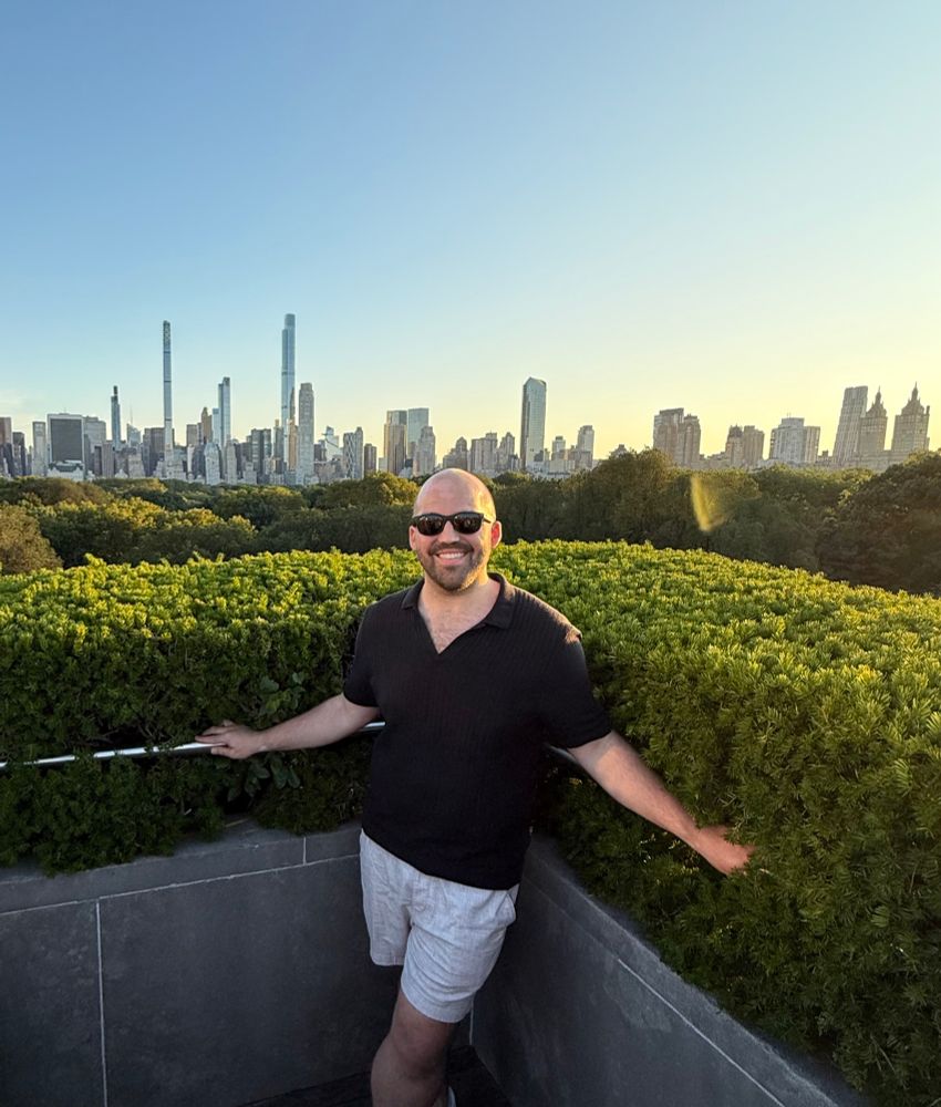 op standing on a balcony in a black knit polo, beige linen shorts, and black ray bans standing on the rooftop of the met