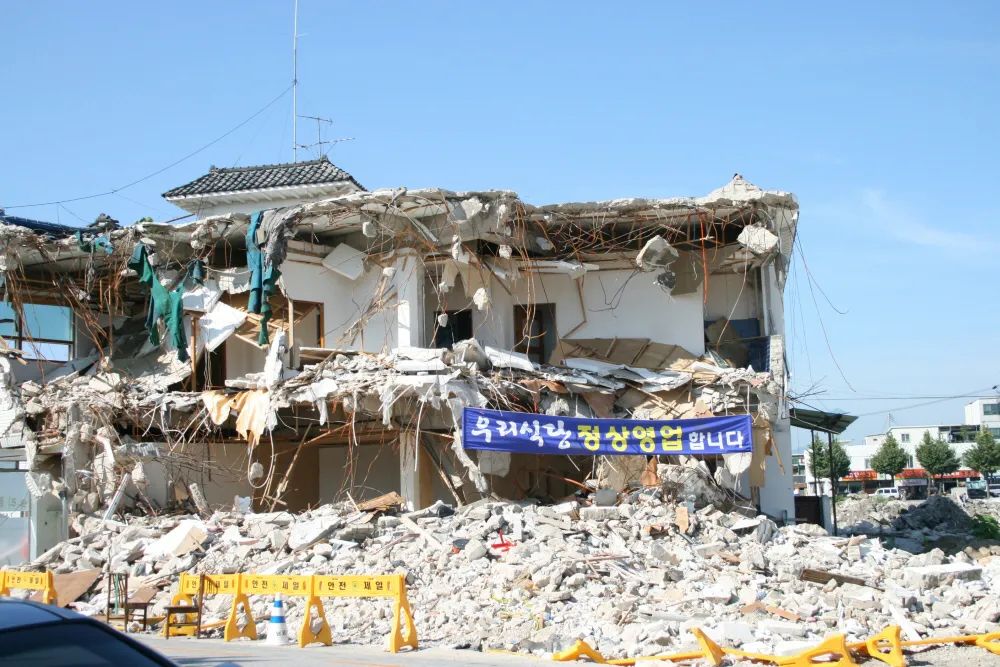 Un bâtiment de 2 étages à moitié démoli de manière transversale, révélant ainsi certaines des pièces, avec une pile de gravats devant, et une banderole bleue accrochée au niveau du rez-de-chaussée.  