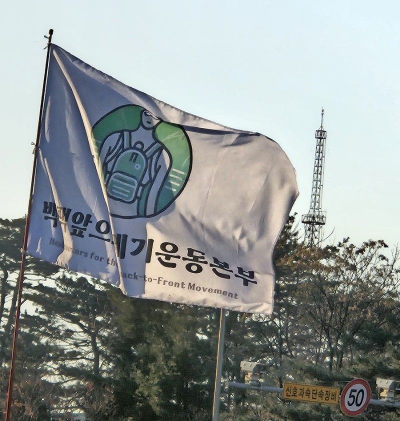 Drapeau blanc flottant au vent, avec un texte en coréen et une illustration en noir, blanc et vert montrant une personne avec un sac-à-dos porté à l'avant.