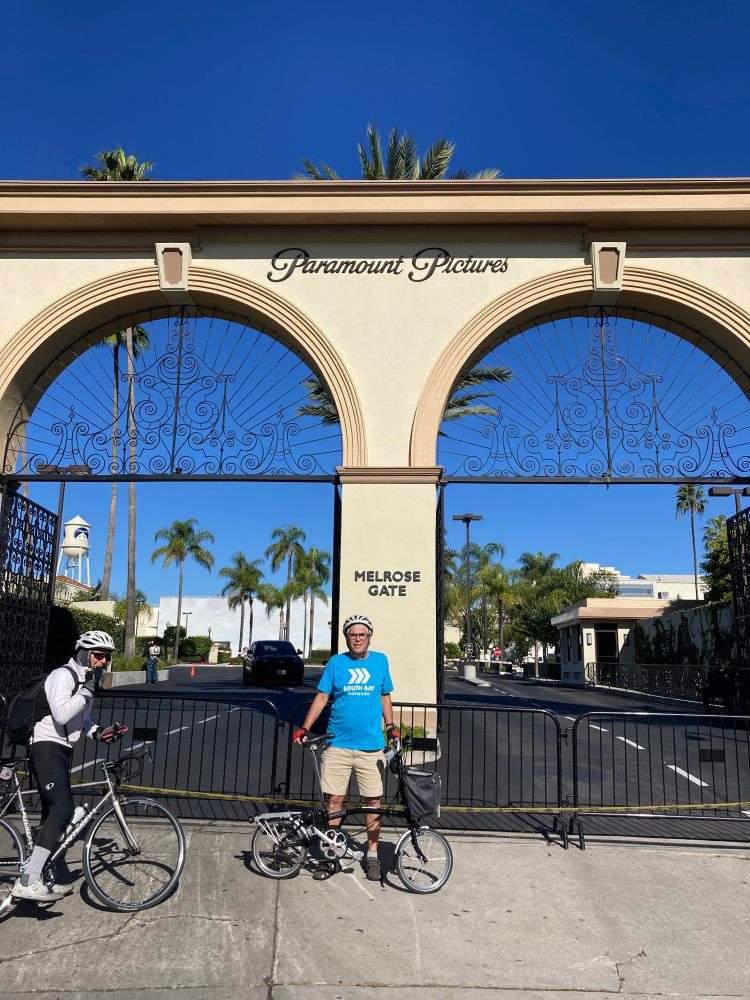 Paramount Pictures double arch and iron gate. A guy with a turquoise blue South Bay Forward shirt poses in front with his bike