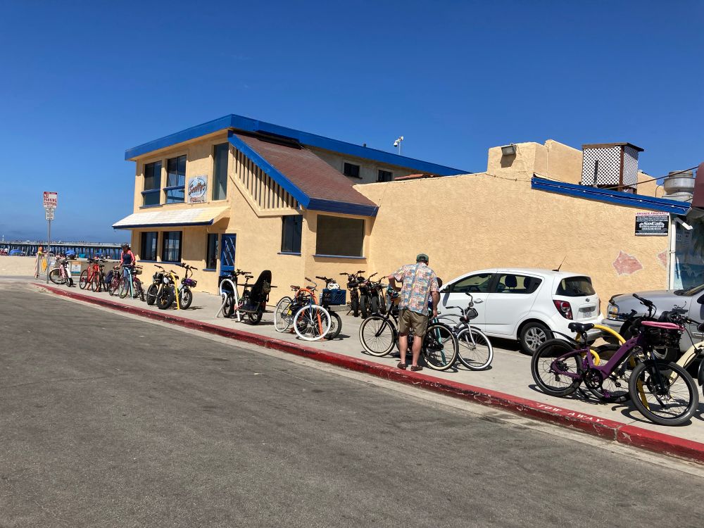 Tan stucco building next to beach bike path. Staple racks line the sidewalk. Each has 2 bikes locked to it. 2 cars in a tandem parking spot and 3 bikes in front of the cars. 