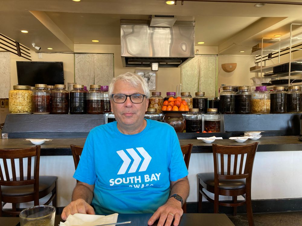 An with glasses, white hair and blue/white South Bay Forward Tshirt sitting at a table in front of bar seating. A shelf above the bar is lined with jars of special liquors and a pile of ripening orange Hachiya persimmons