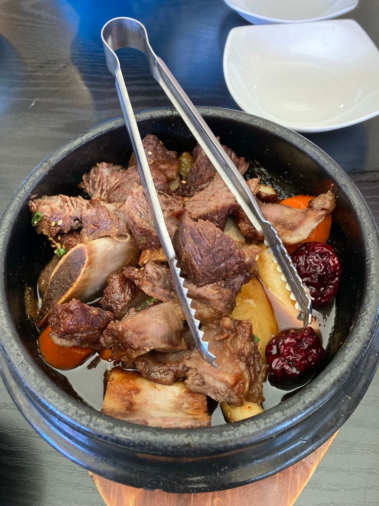 Rib stew in black stone pot and steel tongs. Red jujubes, white turnip and rice sticks, orange carrots, brown portobello mushrooms and beef ribs