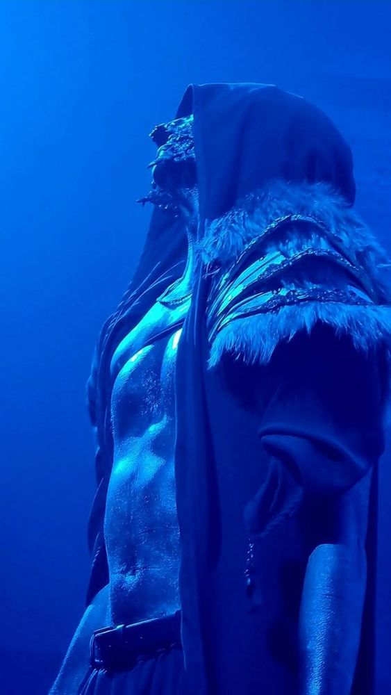 Vessel from Sleep Token in blue lighting wearing a black cloak revealing his very lickable abdomen 