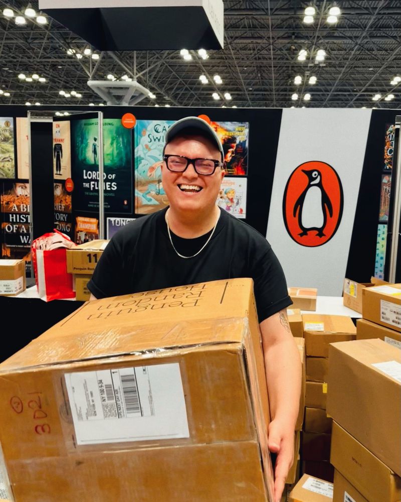 Me, wearing a black tshirt holding a large box in front of the Penguin Random House Comic Con booth.