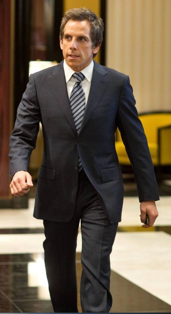 Ben Stiller in a gray suit and striped tie walking through a marble lobby.