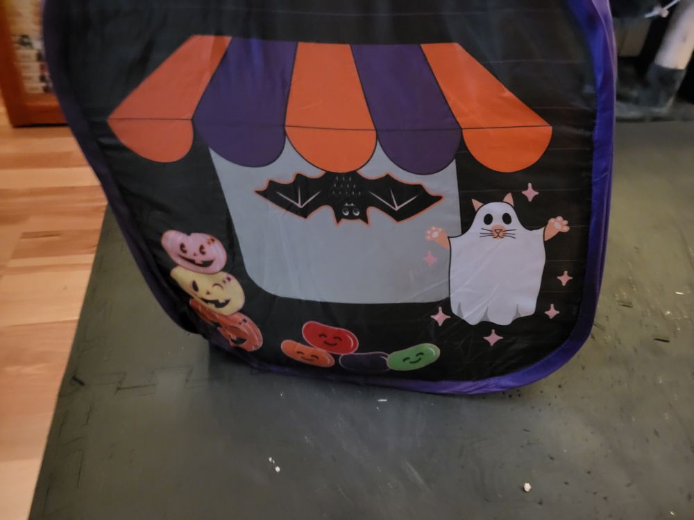 One side of the pop up cat candy store. It is black with a printed on window under an orange and purple overhang with a black bat hanging from it with its wings spread. To the right is a ghost cat, under eath are smiley jelly beans, and to the left a tower of pumpkins (orange on the bottom, yellow in the middle, pink on top).