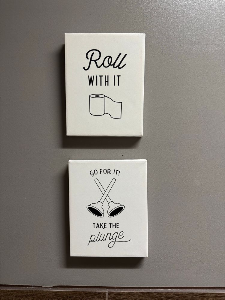 Two pictures on a grey wall. One reads “roll with it” and has an image of a roll of toilet paper; the other says “go for it! Take the plunge” and depicts two dancing toilet plungers. 