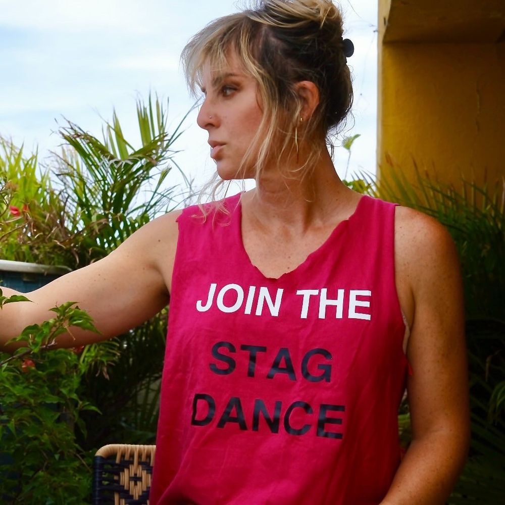 A photo of Torrey Peters looking off to the left of the shot, wearing a pink t-shirt that has been cut at the neck and sleeves to make a tank top. The t-shirt reads, 'JOIN THE STAG DANCE'. 