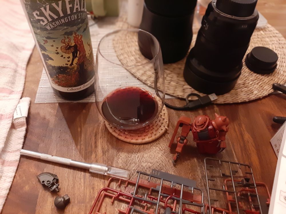 A pic of a delicious bottle of Washington state red opened with a partially sipped glass among camera and model building hobbies on a wood table.