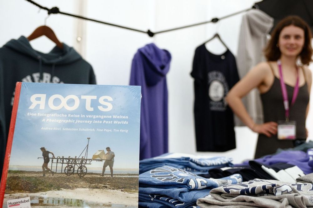 In einem Zelt hängen und liegen Kleidungsstücke. Davor steht ein großformatiges Buch mit dem Titel "ROOTS". Im Hintergrund eine steht eine Frau mit den Händen in den Hüften 