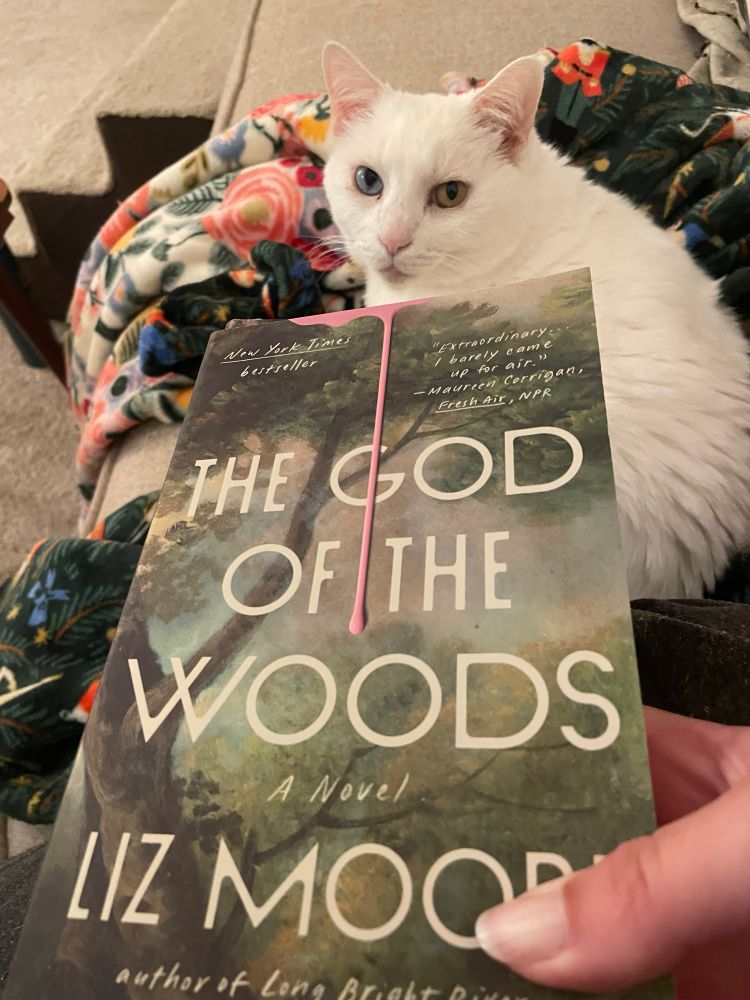Me holding paperback edition of The God of the Woods by Liz Moore in front of my white cat. 
