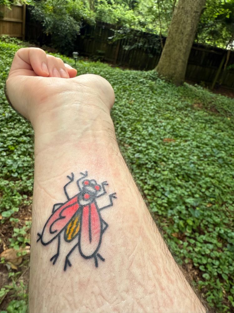 My arm with a tattoo of a fly, the eyes and wings are red and a little yellow on the back. Behind my arm is part of our backyard filled with clovers, a tree, and some vines on a fence.