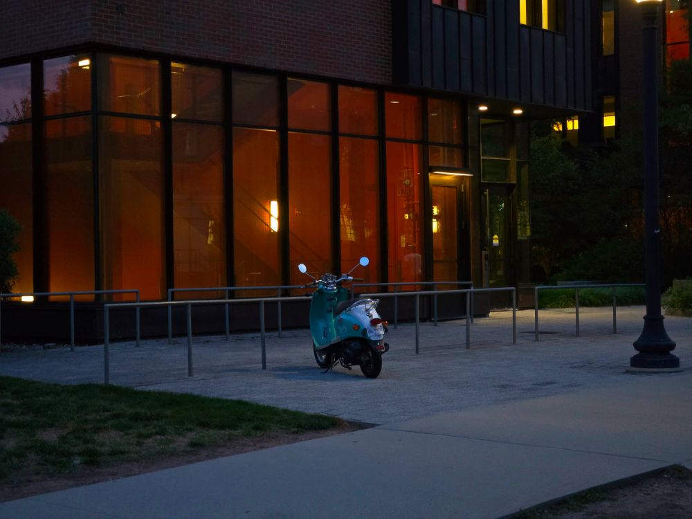 An old scooter next to a warmly lit modern building.