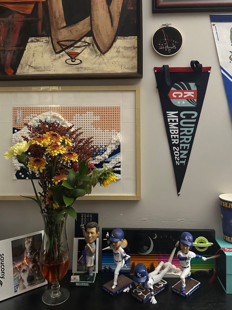 Photo of a table with a picture frame of two dogs, three Kansas City Royals and a Sporting KC bobbleheads, and a background wall festooned with LEGO Hokusai’s “Great Wave,” a KC Current pennant flag, and a cross-stitch of the infamous KC Chiefs “Jet Chip Wasp” play drawn up. On the table also sits a Pat O’Brien’s hurricane glass as a makeshift vase with assorted red and yellow flowers (carnations, sunflowers, etc.) in it.