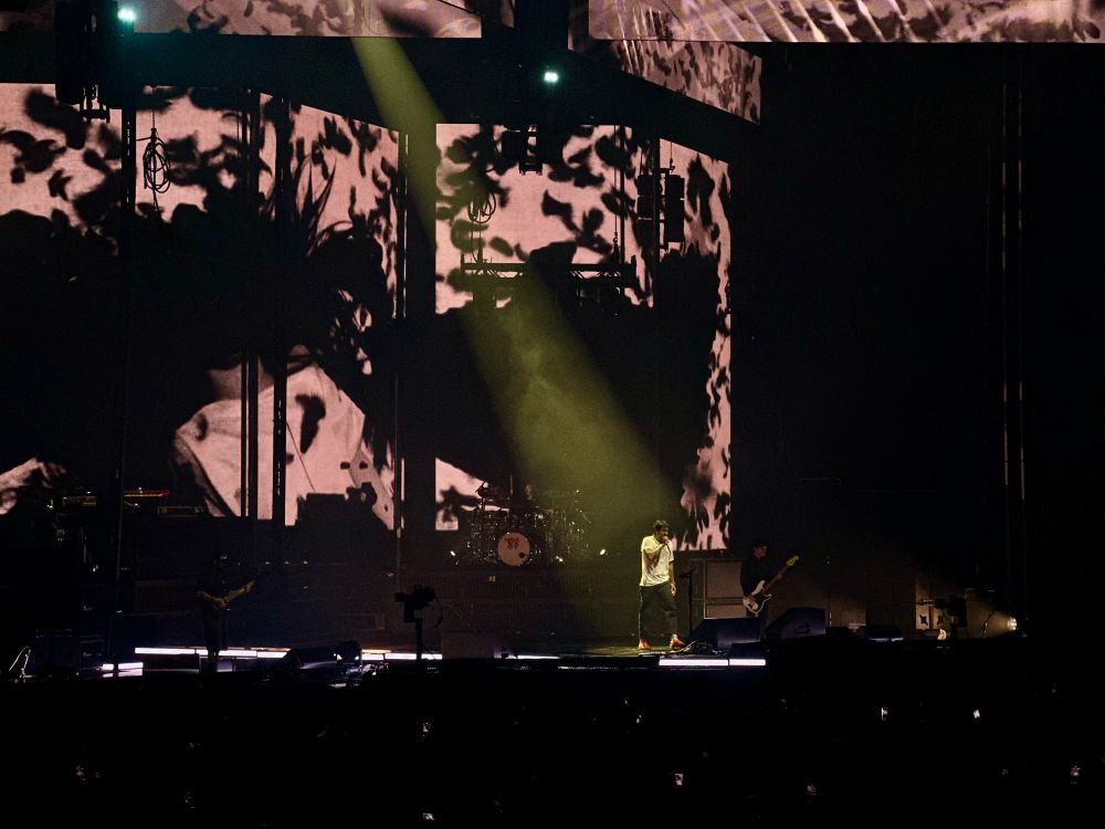 Low-exposure photo of Chino Moreno, lead singer of deftones, walking on stage with a microphone to his mouth in a hazy yellow spotlight at T-Mobile Center in Kansas City, MO on September 17, 2025.