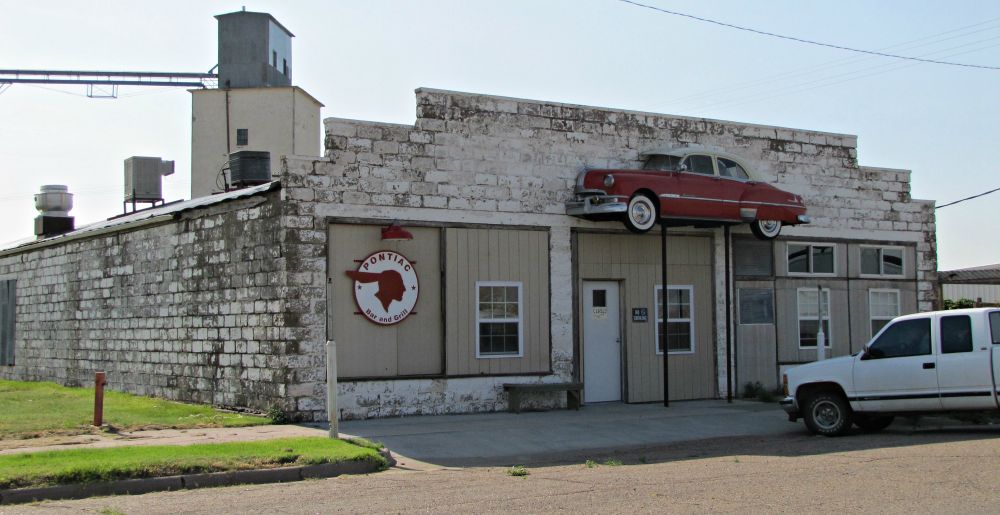 Photo of an old white limestone brick building with half of a 1950s Pontiac coupe mounted on the front of the building over the door.