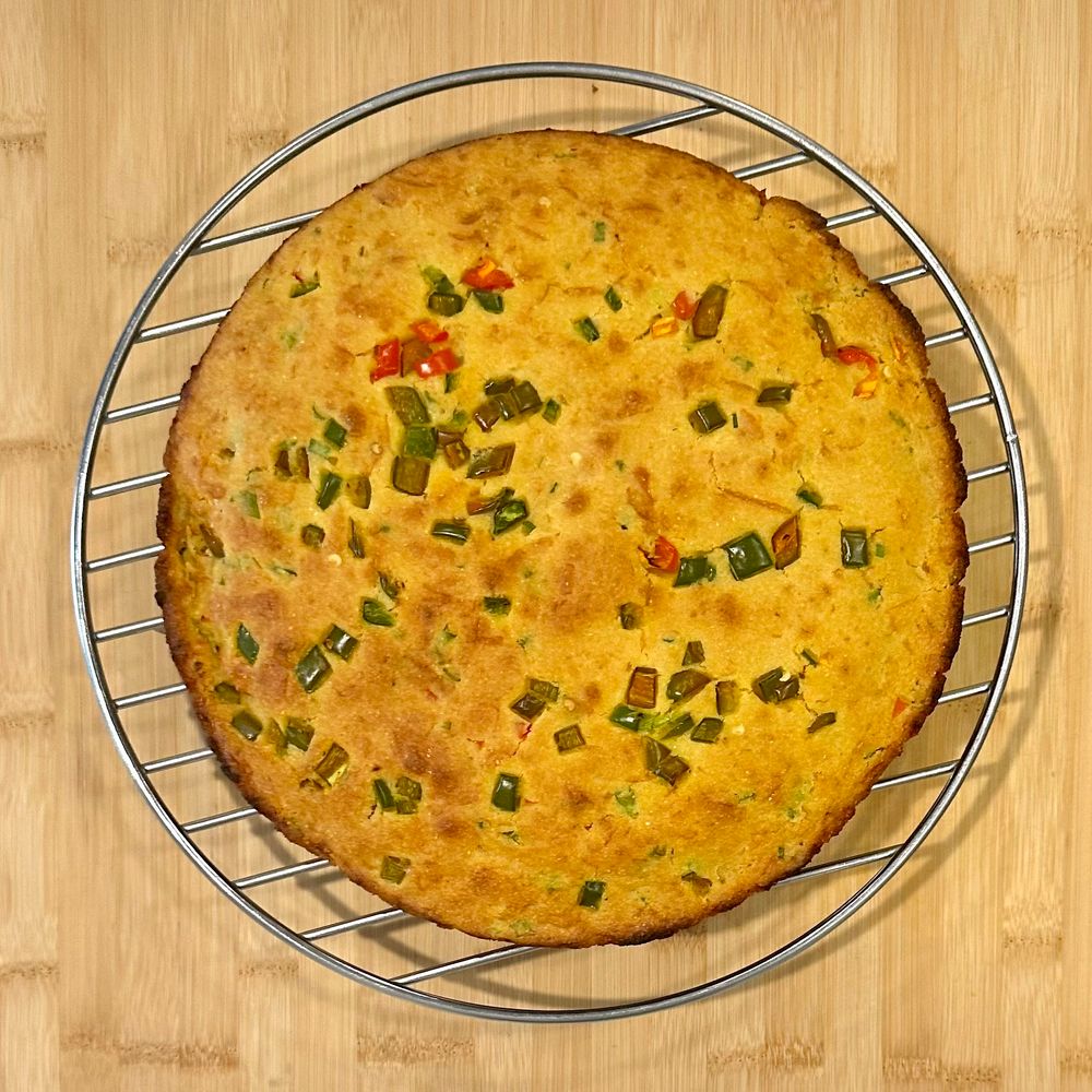 Overhead view of circular jalapeño cornbread sitting on a cooling rack. The bread is toasted yellow with brown crusted edges of the circle, and diced fresh jalapeños cooked into the top layer.