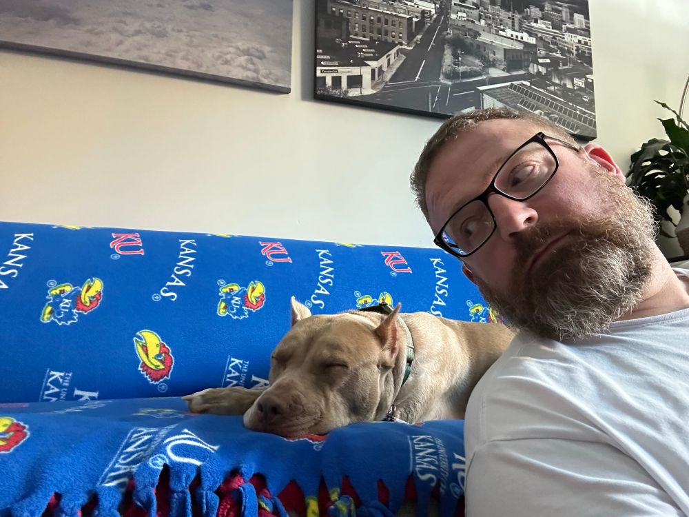 Nala, the brown pittie, laying down with eyes closed on a couch with a KU Jayhawks throw, as the author sits on the floor looking over his right shoulder at her.