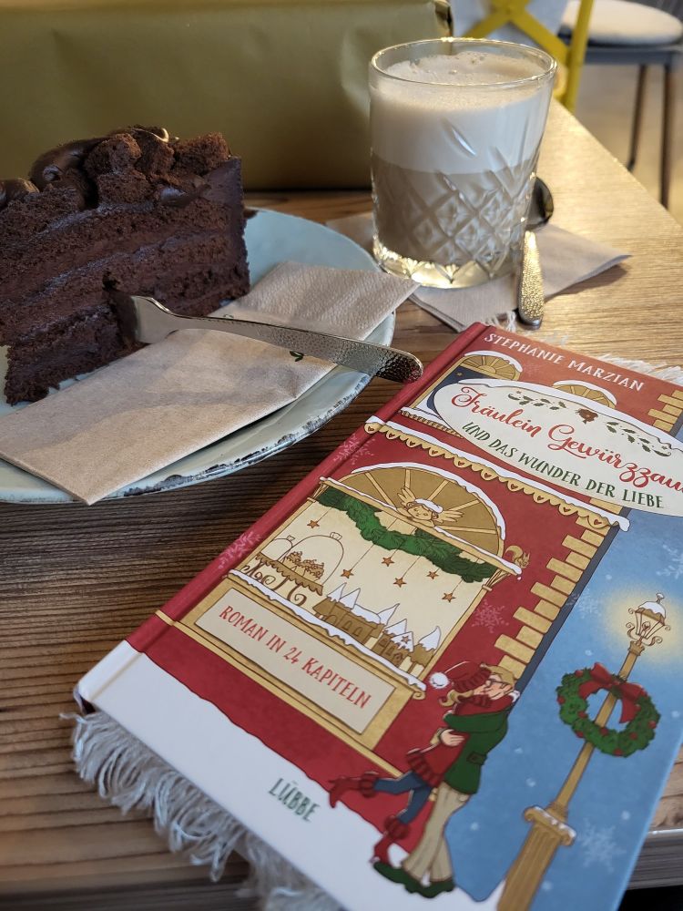 Ein rotes Buch liegt auf einem Tisch. Dahinter steht ein Stück schokoladenkuchen und ein Glas mit Kaffee