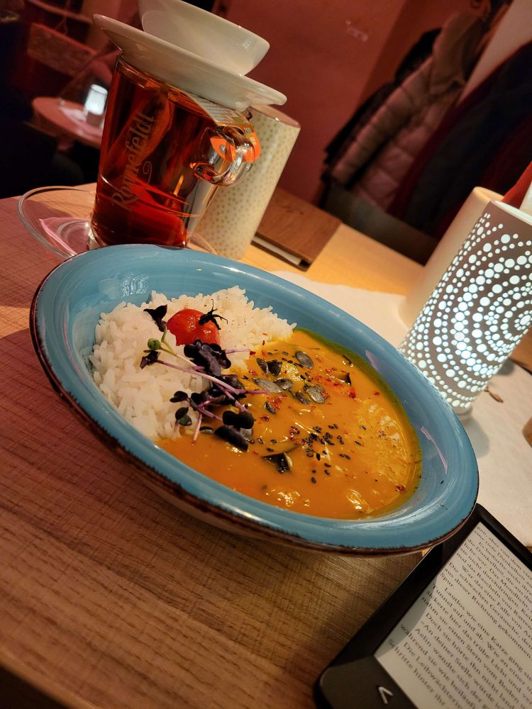 Kürbis curry mit reis in einem blauen teller, daneben steht ein Glas tee auf dem tisch und ein ereader liegt dazu