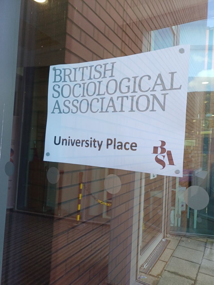 A picture of a piece of paper stuck to a window.

The paper says 'British Sociologocal Association, University Place.'

On the bottom right of the paper is the logo for the British Sociological Association. 