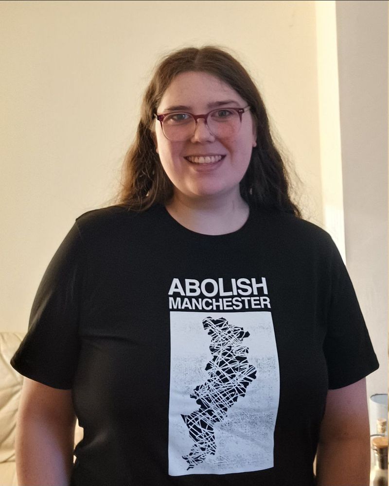 Me (Sylvia), standing up behind a white background, wearing a black t-shirt that says 'Abolish Manchester' in all caps and white text. Underneath the text, there is a white a graphic of the metropolitan borough of Manchester scribbled over.

The t-shirt is from STAT magazine.