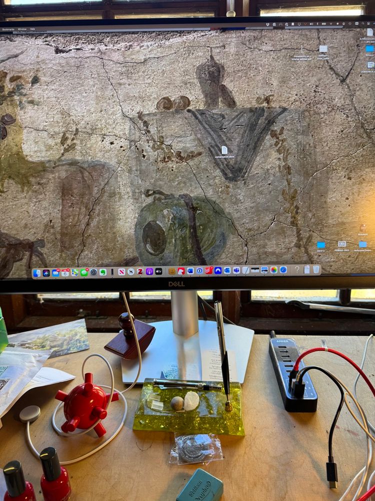 Wooden desk with assorted cables, a roller blotter, knickknacks and tchotchkes, including a green acrylic pen rest. Computer screen sits on desk and fills most of the image with a closeup of a still life fresco (muted colours) as the wallpaper. 