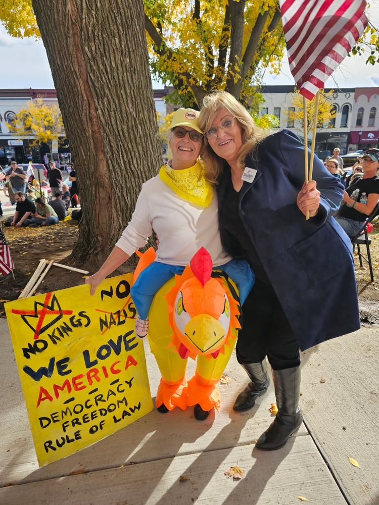 Protester is dressed in a chicken.costumee making fun of TACO Trump and Congress's Republican cowards. The homemade sign she's carrying reads: No Kings, No Nazis, We Love America - Democracy, Freedom,& Rule of Law.  Lisa White stands besise her. She is a candidate for WI State Senator.