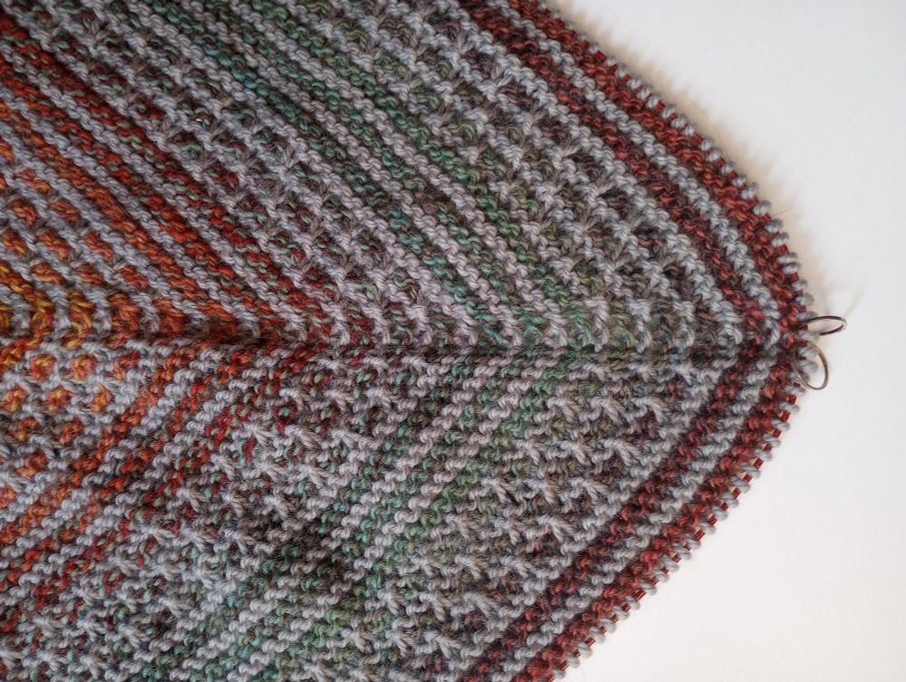 A knitted wrap showing a gradient yarn alternated with a gray yarn. The center has v shaped stripes due to the way the yarn increases slant inwards towards the stitch marker.
