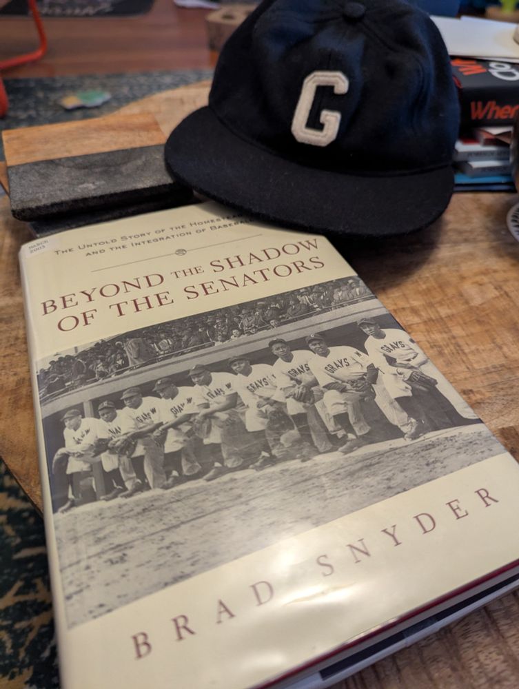 A book titled, "Beyond the Shadow of the Senators by Brad Snyder" sits on a wooden table next to a dark blue baseball cap with a white G (a Homestead Grays cap)