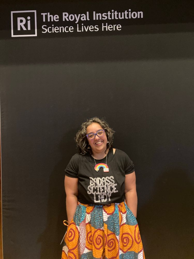 Hana wearing a black tshirt which says badass science lady, a rainbow pencil necklace, traditional Indian earrings and a brightly patterned skirt in oranges and blues

The royal institution logo and motto is above her 