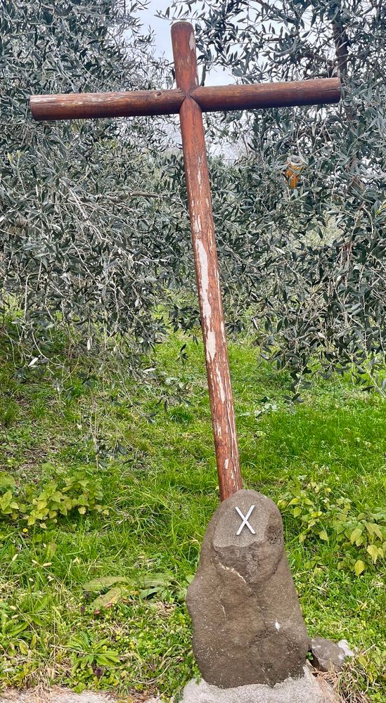 Ein Kreuz am Wegesrand darunter ein Stein mit einem X drauf. Olivenbäume im Hintergrund. 