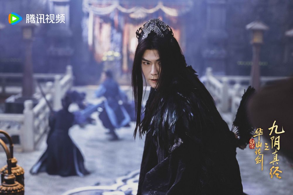 promo image showing ji chen with long black hair, impressive winged eyeliner, a dark metal crown, a dark robe with exaggerated shoulders covered in black feathers. he appears to be in the middle of a battle and his hair is falling in front of his eyes 