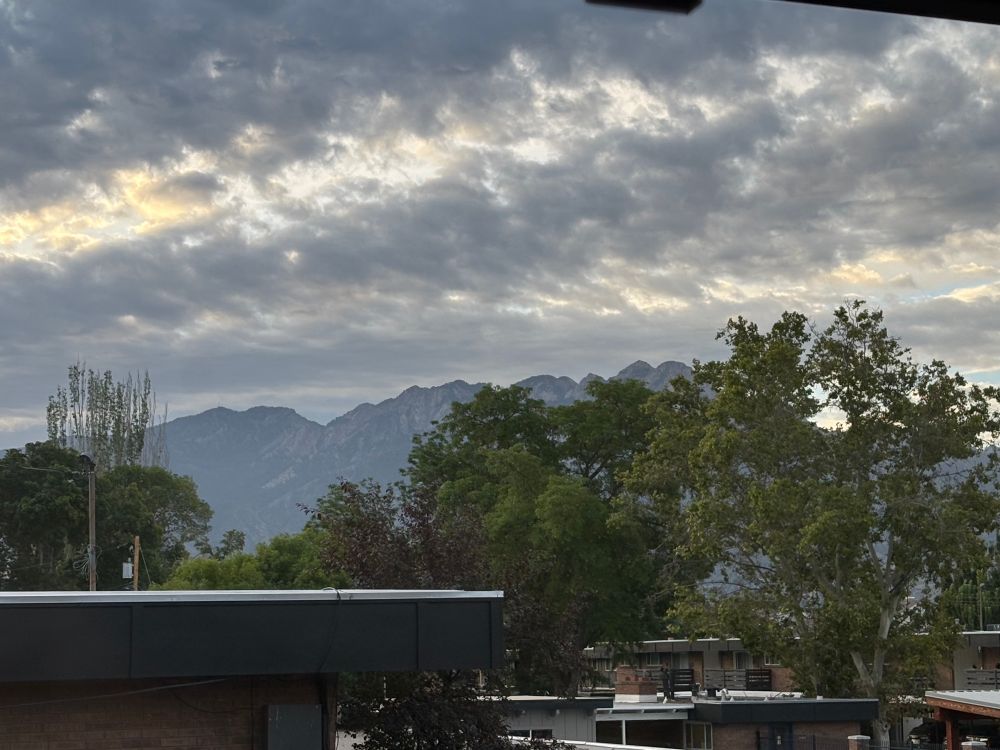 Skittery grey clouds with light peeking through and some golden light. Mountains in the mist ? Very green leafy trees. Bits is apartment complex at the bottom. 