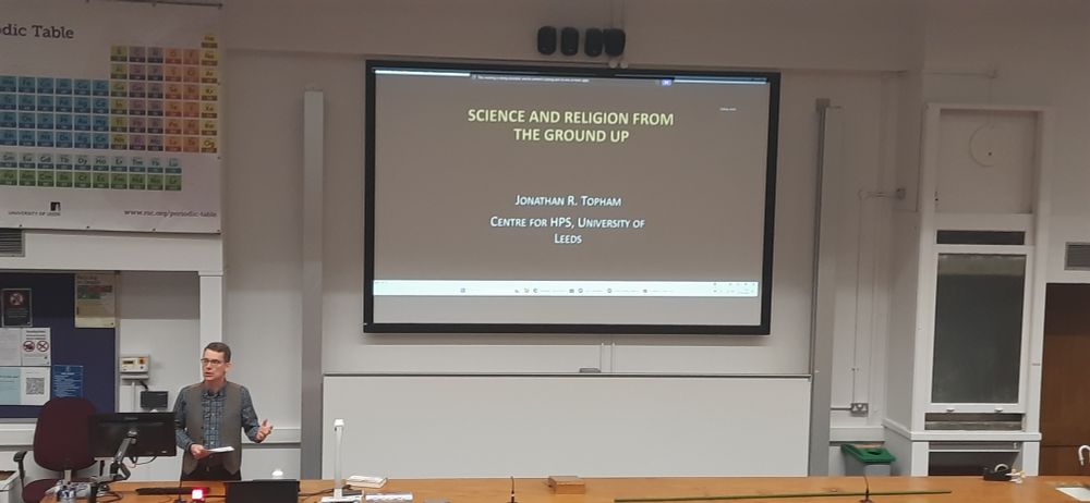 Image of Jon Topham standing to the left of a screen with "Science and Religion from the Ground Up" on it centre front. And a periodic table in the background on the left.
