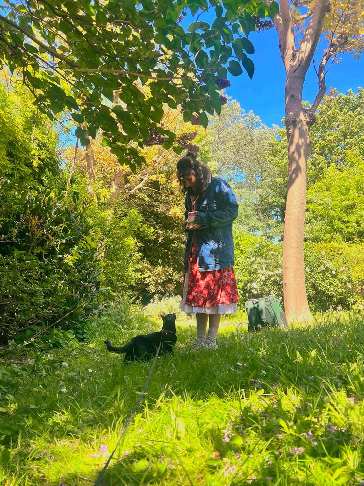 Woman (in a red skirt, white petticoat and large jeans jacket) and a black cat in a sun kissed and lusciously flowering garden. They're both standing, the woman looking down at the cat and the cat, tail up, looking up at the woman.