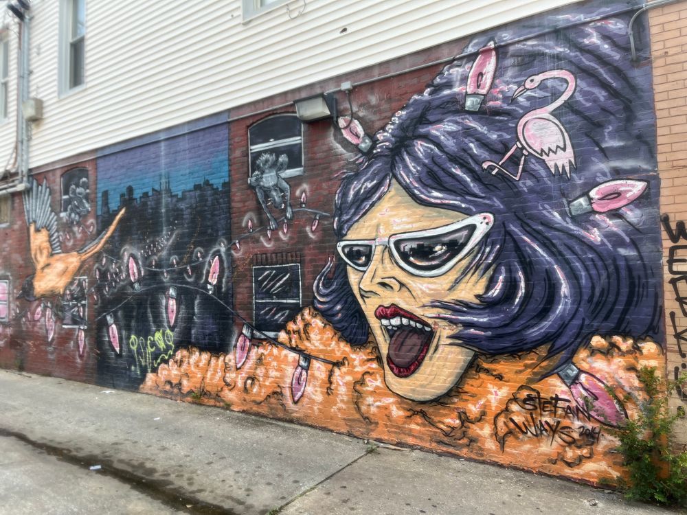 Purple haired woman in graffiti with white glasses and light bulbs and flamingos in her hair. 