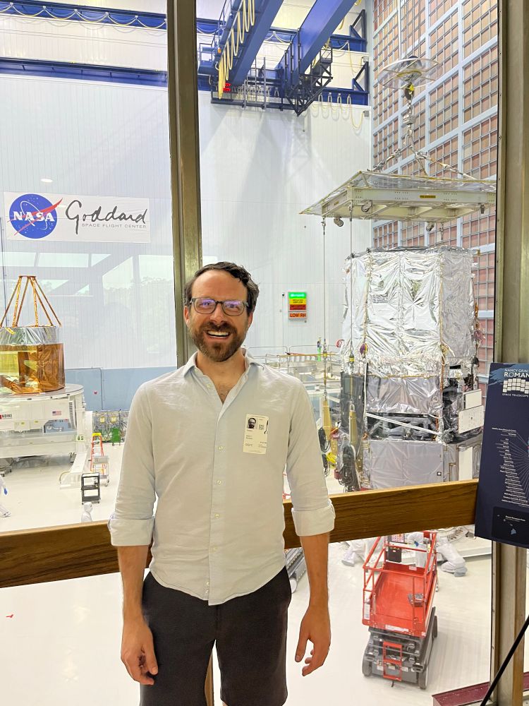 Brett standing in front of a window overlooking the big clean room at NASA Goddard, where spacecraft are assembled. The Roman Space Telescope is behind Brett to the right. 