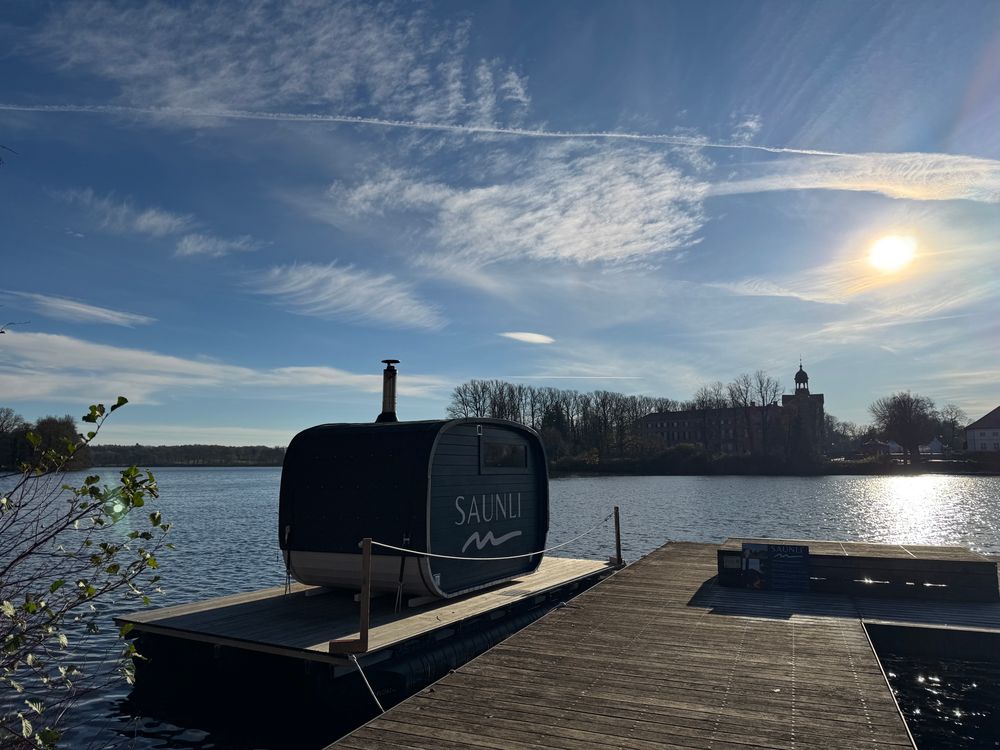 Gegenlicht Aufnahme 
Ein Holzsteg auf dem Eutiner See mit einem kleinen Ponton, worauf eine kleine Sauna Kabine steht. Im Hintergrund das Schloss 