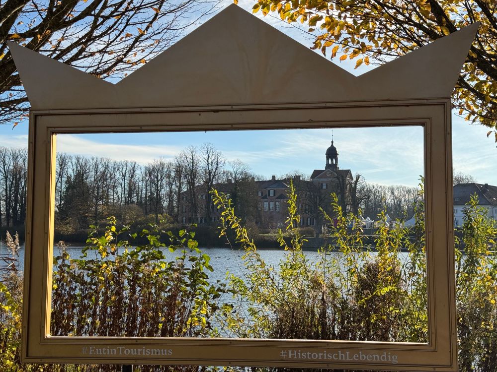 Ein leerer Bilderrahmen vor Büschen und See, dahinter das Eutiner Schloss (Museum)