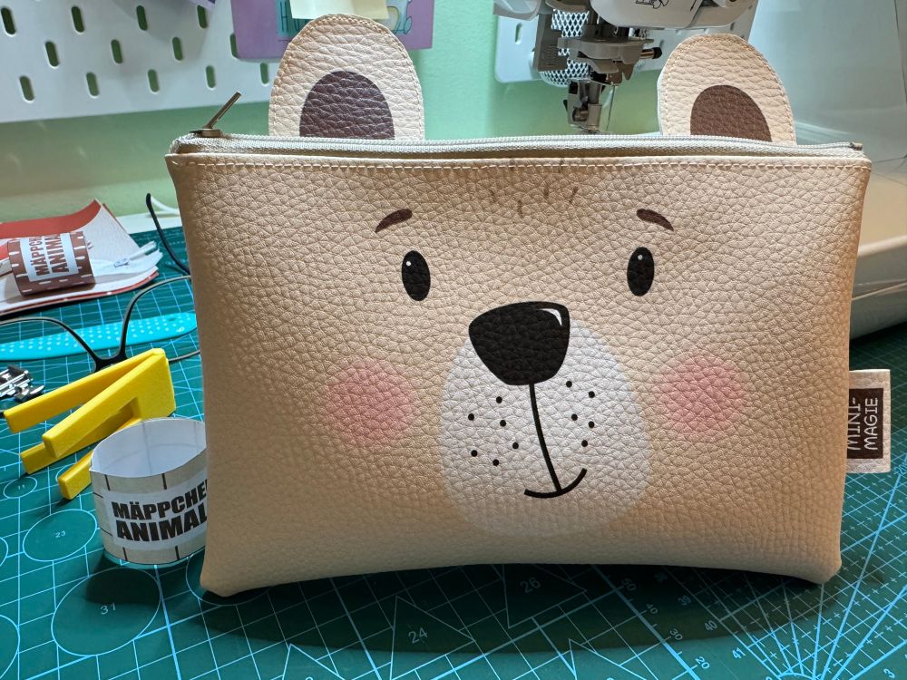 Auf dem Nähtisch steht an die Nähmaschine gelehnt eine Kunstleder Mäppchen im Bärchen (Kopf) Design. Drumherum stehen ein Zettel (Mäppchen Animals) ein Hebamme (Höhenausgleich beim nähen) eine Brille, ein Wendetool, Zuschnitt des nächsten Mäppchens. 