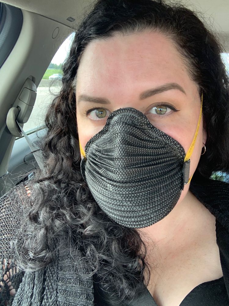 A selfie of Elisabeth, who is a white person with long, dark brown curly hair. She is sitting in her car and wearing a black respirator with yellow straps (Moldex M4620), a black cardigan, and a black dress.