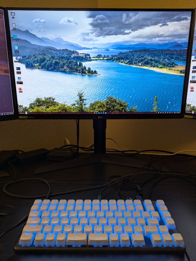 A picture of my desktop with my monitor at the top and my keyboard at the bottom. The monitor is showing the COSMIC desktop environment with a colorful background. The keyboard has LEDs that are matched to an accent color extracted from that background, in this case, blue.